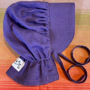 May include: A purple linen bonnet with a gathered crown and a wide brim. The bonnet has two ties that can be tied under the chin. The bonnet has a small label with a dragonfly design and the text "Erica".
