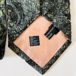 May include: A black and green patterned silk tie with a peach-colored lining. The tie has a black label with the text "SHELL: 100% SILK LINING: 100% ACETATE INTERLINING 60% POLYESTER 40% ACRYLIC" and another black label with the text "Entertainment Inc. SERVED".