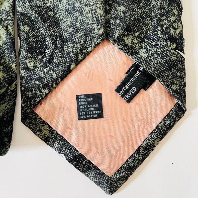 May include: A black and green patterned silk tie with a peach-colored lining. The tie has a black label with the text "SHELL: 100% SILK LINING: 100% ACETATE INTERLINING 60% POLYESTER 40% ACRYLIC" and another black label with the text "Entertainment Inc. SERVED".