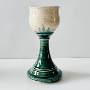 May include: A handmade ceramic goblet with a speckled beige bowl and a dark green stem and base. The goblet has a wide, rounded bowl and a tall, flared stem. The base is also flared, providing stability. The goblet is likely for decorative use.