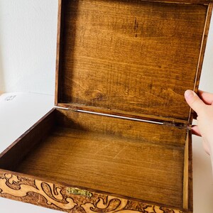 May include: Open wooden box with a hinged lid. The box is stained a warm brown color, with a lighter interior. The front of the box features an ornate, carved design. The box is empty and ready to be filled.