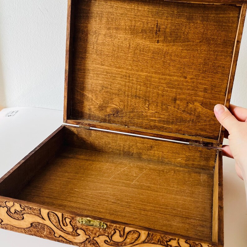 May include: Open wooden box with a hinged lid. The box is stained a warm brown color, with a lighter interior. The front of the box features an ornate, carved design. The box is empty and ready to be filled.