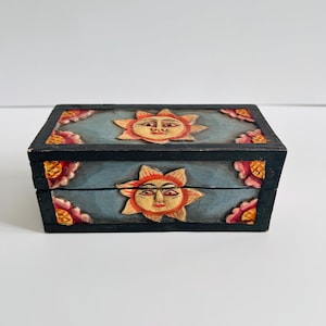 Könnte beinhalten: Kleine, rechteckige Holzbox mit Scharnierdeckel. Die Box ist schwarz lackiert mit hellblauem Deckel und Vorderseite. Ober- und Vorderseite zeigen geschnitzte Sonnengesichter und florale Eckakzente in Orange, Rosa und Gelb.