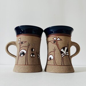 Peut inclure: Deux tasses en céramique avec un motif abstrait brun et blanc. Les tasses ont un glaçage bleu à l'intérieur et un glaçage brun à l'extérieur. Le motif représente des oiseaux stylisés avec des motifs géométriques.