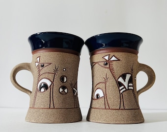 Puede incluir: Dos tazas de cerámica con un diseño abstracto marrón y blanco. Las tazas tienen un esmalte azul en el interior y un esmalte marrón en el exterior. El diseño presenta pájaros estilizados con patrones geométricos.