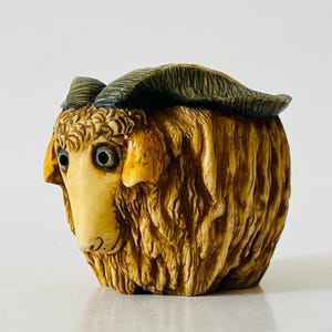 May include: A detailed figurine of a sheep with a cream-colored face and tan fur. The sheep has large, round eyes and dark green horns. The figurine is sculpted with intricate details, giving it a lifelike appearance.