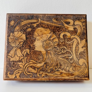 May include: A rectangular wooden box with a detailed pyrography design. The artwork features a woman's profile, floral elements, and flowing lines. The box has a warm, natural wood tone with darker burn marks.