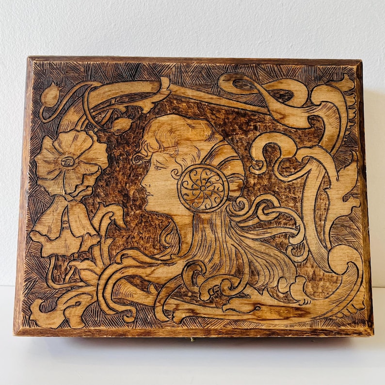 May include: A rectangular wooden box with a detailed pyrography design. The artwork features a woman's profile, floral elements, and flowing lines. The box has a warm, natural wood tone with darker burn marks.