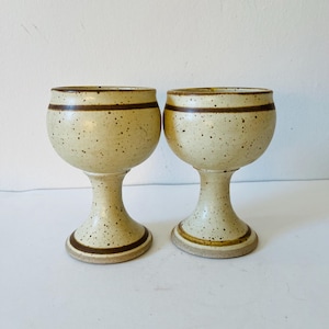 Puede incluir: Dos copas de cerámica beige con manchas y bandas marrones alrededor de la parte superior e inferior del cuenco y la base. Las copas tienen un cuenco redondeado y un tallo alto y delgado.
