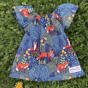 May include: A blue dress with a forest and fox print. The dress features a pattern of orange foxes, yellow flowers, and green foliage on a dark blue background. The dress has flutter sleeves and a gathered neckline.