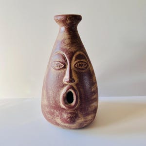 Op de afbeelding: Een bordeauxrode keramische vaas met een gezichtsontwerp. De vaas heeft gebeeldhouwde ogen, een neus en een open mond. De vaas heeft een getextureerd oppervlak en een smalle hals, geschikt voor interieurdecoratie.