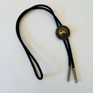 Op de afbeelding: Een zwarte bolo tie met een ronde hanger van zilver en bruin. De hanger heeft een decoratief ontwerp en een bruine steen in het midden.