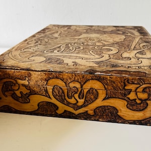 May include: A wooden box with intricate carvings on the lid and sides. The box features a light brown color with darker brown detailing, showcasing a detailed design. The carvings depict a figure and floral patterns.