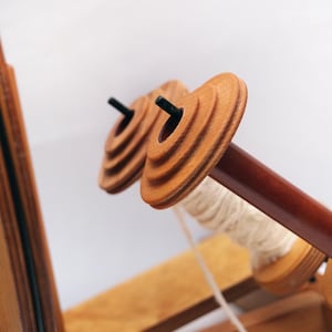 May include: Close-up of a wooden weaving loom with two spools in focus. The spools are light brown with a dark brown centre rod. White thread is wrapped around one spool. The loom is made of light brown wood.