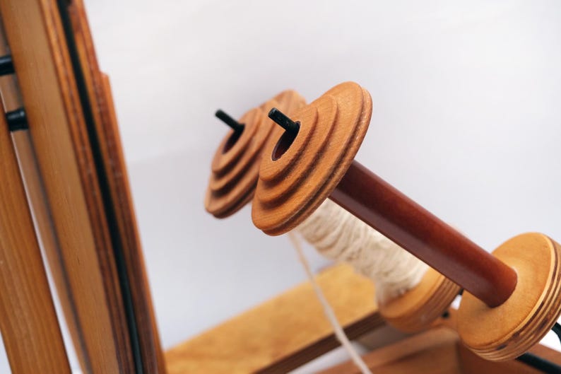 May include: Close-up of a wooden weaving loom with two spools in focus. The spools are light brown with a dark brown centre rod. White thread is wrapped around one spool. The loom is made of light brown wood.