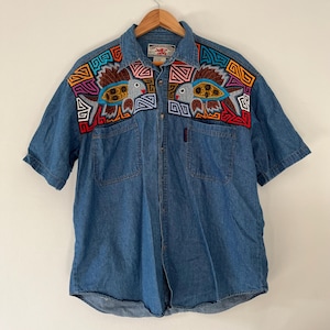 Op de afbeelding: Een blauw spijkeroverhemd met korte mouwen. Het overhemd heeft geborduurde visontwerpen in bruin, rood, geel en blauw op de borst. Het heeft twee voorzakken en een klein rood label. Het overhemd is dichtgeknoopt.