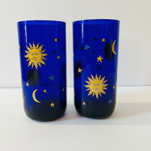 May include: Two cylindrical cobalt blue glass tumblers featuring a repeating celestial pattern. The design includes yellow sun faces with rays, golden crescent moons, and various small stars in yellow and light blue scattered across the surface.
