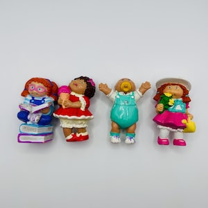 May include: Four vintage Cabbage Patch Kids figurines. One is reading on a stack of books, another holding an ice cream cone, a third with arms raised, and the last holding flowers and a purse. Each figurine is brightly colored.