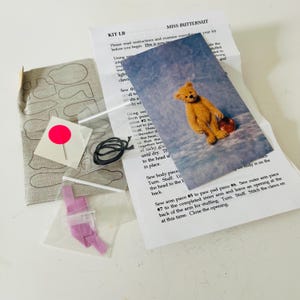 May include: A craft kit with instructions and materials for making a small teddy bear. The kit includes printed instructions, fabric pieces, a needle, thread, and a picture of the finished bear. The bear is holding a small red object.