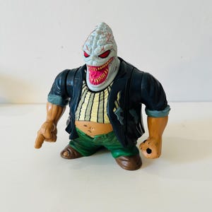 Peut inclure: Figurine détaillée d'un monstre avec une tête pâle, des yeux rouges et une grande bouche dentée. Il porte une veste noire sur une chemise rayée, un pantalon vert et des chaussures marron. La figurine est posée avec un bras tendu.