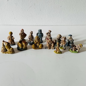 Peut inclure: Une collection de quatorze petites figurines en céramique peintes à la main représentant divers personnages et objets, dont une maison en forme de chaussure, une personne en cape rouge, un personnage avec des poids et des tenues historiques.