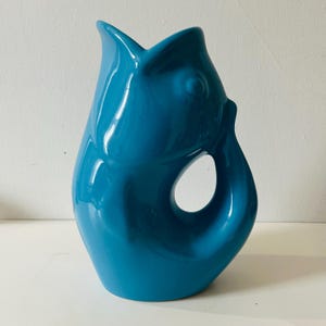 May include: A turquoise ceramic jug in the shape of a fish. The jug has a large opening on the side and a flared top. The design is smooth and modern, with a high-gloss finish. The jug is a decorative item, suitable for display.