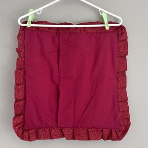 May include: A burgundy pillowcase with a ruffled edge. The pillowcase is a solid burgundy color in the center, with a patterned border of burgundy fabric with small white polka dots. The pillowcase is hanging on a white hanger.