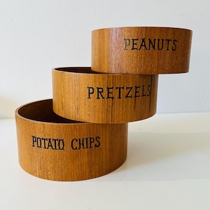 May include: Three wooden snack bowls stacked on top of each other. The top bowl reads "PEANUTS", the middle bowl reads "PRETZELS", and the bottom bowl reads "POTATO CHIPS". The bowls are made of a light brown wood.