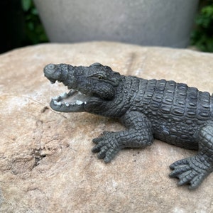 Peut inclure: Une figurine de crocodile gris foncé avec la bouche ouverte, montrant des dents blanches. La figurine est posée sur un rocher gris clair.