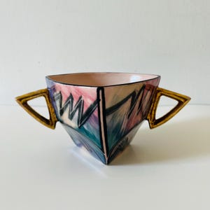 Puede incluir: Un cuenco triangular de cerámica con dos asas angulares en tono dorado. El exterior presenta un patrón abstracto pintado a mano con pinceladas en rosa, azul verdoso y morado, acentuado por líneas negras que definen la forma geométrica.