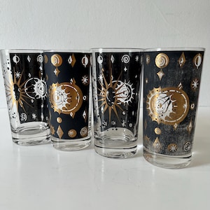 May include: A set of four clear glass tumblers featuring a black background with gold and white celestial motifs, including suns, moons, stars, and geometric shapes. Each cylindrical glass has a flat base and a smooth rim for drinking.