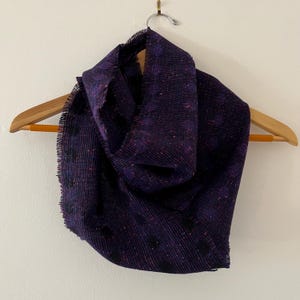 May include: A purple and black patterned scarf with a fringed edge. The scarf is hanging on a wooden hanger.