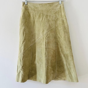 Può includere: Una gonna in camoscio verde oliva chiaro con vita alta, cinturino strutturato e dettagli con cuciture diagonali che creano un effetto a pannelli sul davanti. La gonna presenta una silhouette svasata e una texture liscia e opaca.
