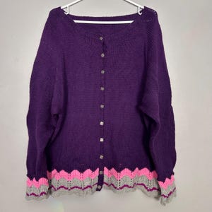 May include: A purple cardigan sweater with a round neckline and long sleeves. The sweater has a button-up front with silver buttons. The bottom hem and sleeve cuffs feature a decorative pattern in pink, gray, and purple.
