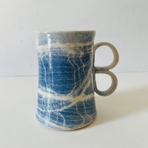 May include: A ceramic mug featuring a speckled blue and white marbled glaze pattern. The cylindrical body has a slightly tapered shape and includes a unique handle design consisting of two stacked, circular loops attached to the side of the mug.