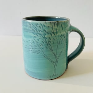 May include: A ceramic mug with a glossy turquoise glaze featuring a hand-painted dark grey tree silhouette on the exterior. The mug has a cylindrical body, a smooth rim, and a curved handle on the right side, showcasing a blend of light and dark blue tones.