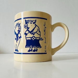 Può includere: Tazza in ceramica color crema con illustrazioni di maiali blu a fumetti. La tazza ha un manico curvo e una fascia blu. Il testo "© 1979 Janet Nottingham" è stampato sul lato. Un oggetto divertente per la cucina.