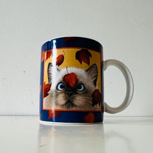 May include: A ceramic coffee mug featuring a cat with blue eyes and autumn leaves. The mug has a blue rim and handle, with a yellow background behind the cat's face. The leaves are red and brown.