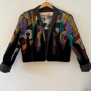 Puede incluir: Una chaqueta de terciopelo negro con detalles de patchwork de colores. La chaqueta tiene una cremallera y un cuello de pie. El texto "Coleccion Judith Ret" es visible en el interior de la chaqueta.