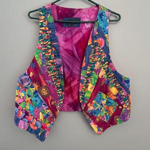 May include: A sleeveless waistcoat featuring a vibrant patchwork design with multicoloured patterns including suns, fish, and floral motifs. The interior lining is a solid magenta fabric, and the garment is displayed on a dark green plastic hanger.