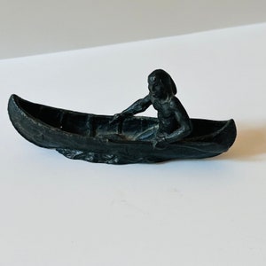 May include: A black figurine of a person paddling a canoe. The canoe is carved with details and the person is in a seated position.