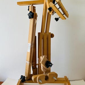 Puede incluir: Un soporte de artesanía de madera ajustable con múltiples componentes giratorios y de bloqueo. El soporte presenta un acabado de madera natural, perillas negras y una base robusta. Está diseñado para sujetar y colocar objetos para manualidades o exhibición.