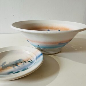 Peut inclure: Ensemble d'un bol et d'une petite assiette en céramique blanche. Le bol a un large bord et est décoré de bandes horizontales roses, bleues et oranges. L'assiette a un motif similaire avec un motif de paysage. Le bol a plusieurs trous.