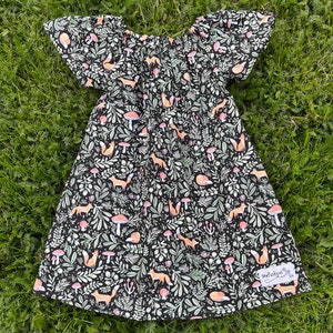 Puede incluir: Un vestido negro con cuello fruncido y mangas cortas abullonadas. El vestido está cubierto con un patrón repetido de zorros, setas y hojas. El vestido tiene una pequeña etiqueta blanca cosida en la parte inferior derecha que dice "SewFancyCo."
