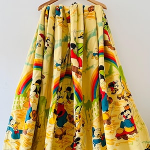 Puede incluir: Manta vintage amarilla con Mickey Mouse, Donald Duck, Goofy y Pluto en monopatines, con arcoíris y un diseño de valla blanca. La manta está colgada de una percha de madera.