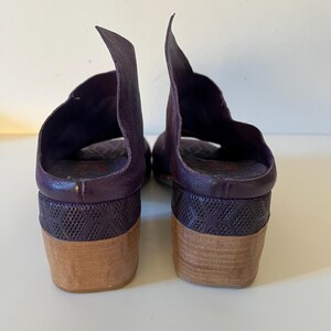 May include: A pair of purple leather sandals with a unique design. The sandals feature a textured heel and a wooden sole. The upper part of the sandal has a distinctive, curved shape. The sandals are set against a white background.