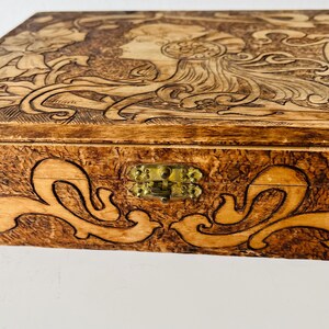 May include: Wooden box with intricate carved design. The lid features a profile of a woman with flowing hair and floral elements. The box has a brass clasp and decorative scrollwork. The wood has a warm, natural tone.