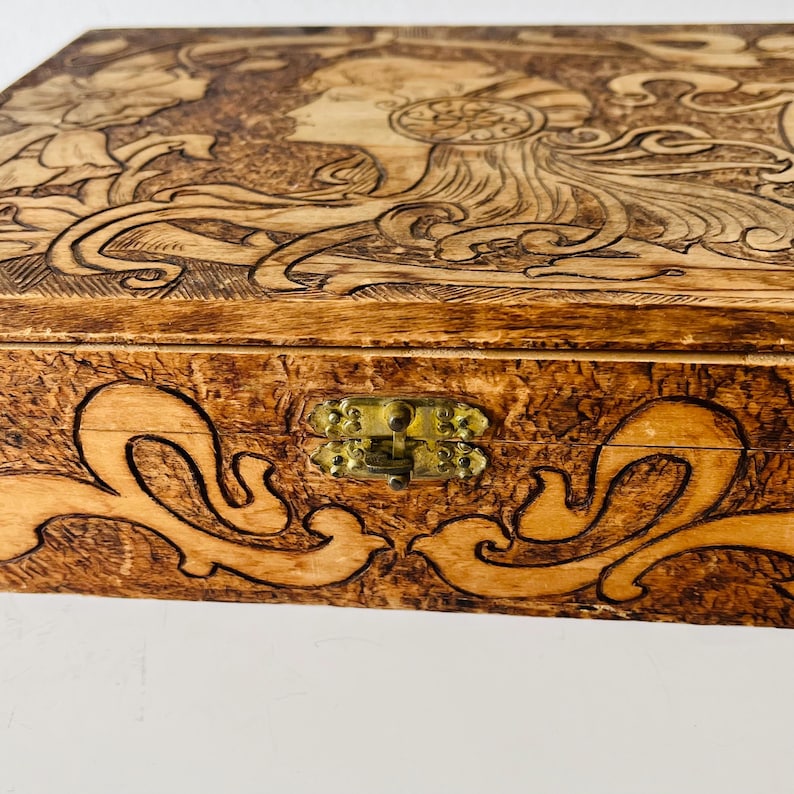 May include: Wooden box with intricate carved design. The lid features a profile of a woman with flowing hair and floral elements. The box has a brass clasp and decorative scrollwork. The wood has a warm, natural tone.