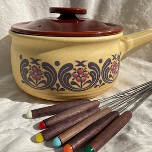 May include: A vintage ceramic fondue pot with a brown lid and floral pattern. The pot is surrounded by six wooden fondue forks with colorful tips.