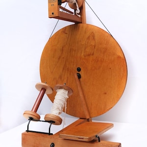 May include: A wooden Lou&euml;t S15 spinning wheel, featuring a large circular wheel and two spindles. The wheel is made of light brown wood and has a small wooden box with the Lou&euml;t logo. Designed for spinning yarn, it is a traditional craft tool.